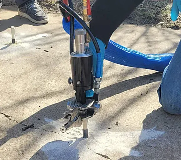 A person in blue gloves operates a pneumatic tool to drill or repair a crack in a concrete surface.
