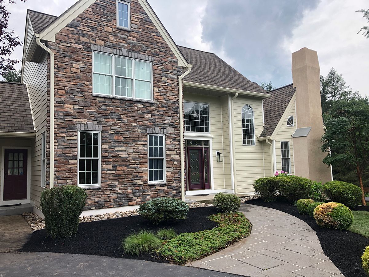 Two-story house with stone facade, beige siding, dark roof, curved walkway, and landscaping under a cloudy sky.