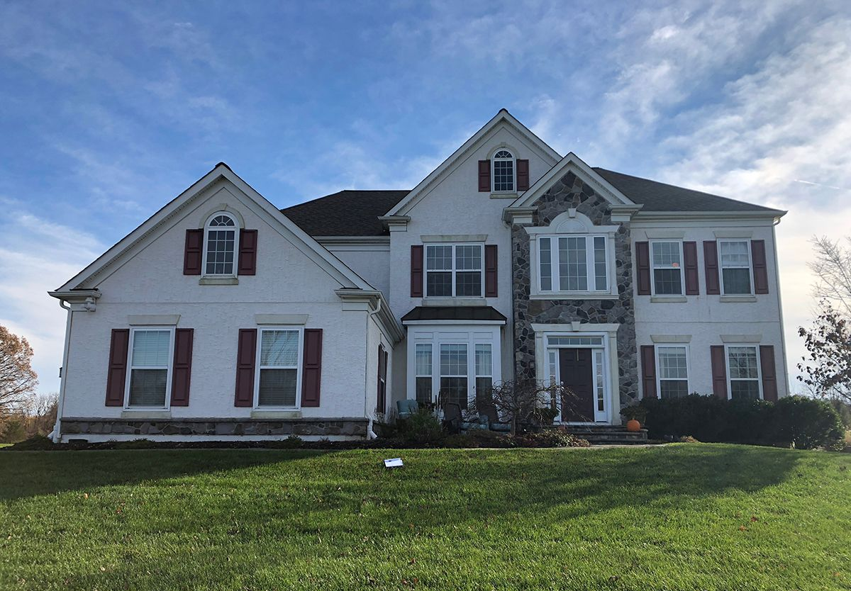 Two-story white house with red shutters and stone accents, set on a green lawn under a blue sky.