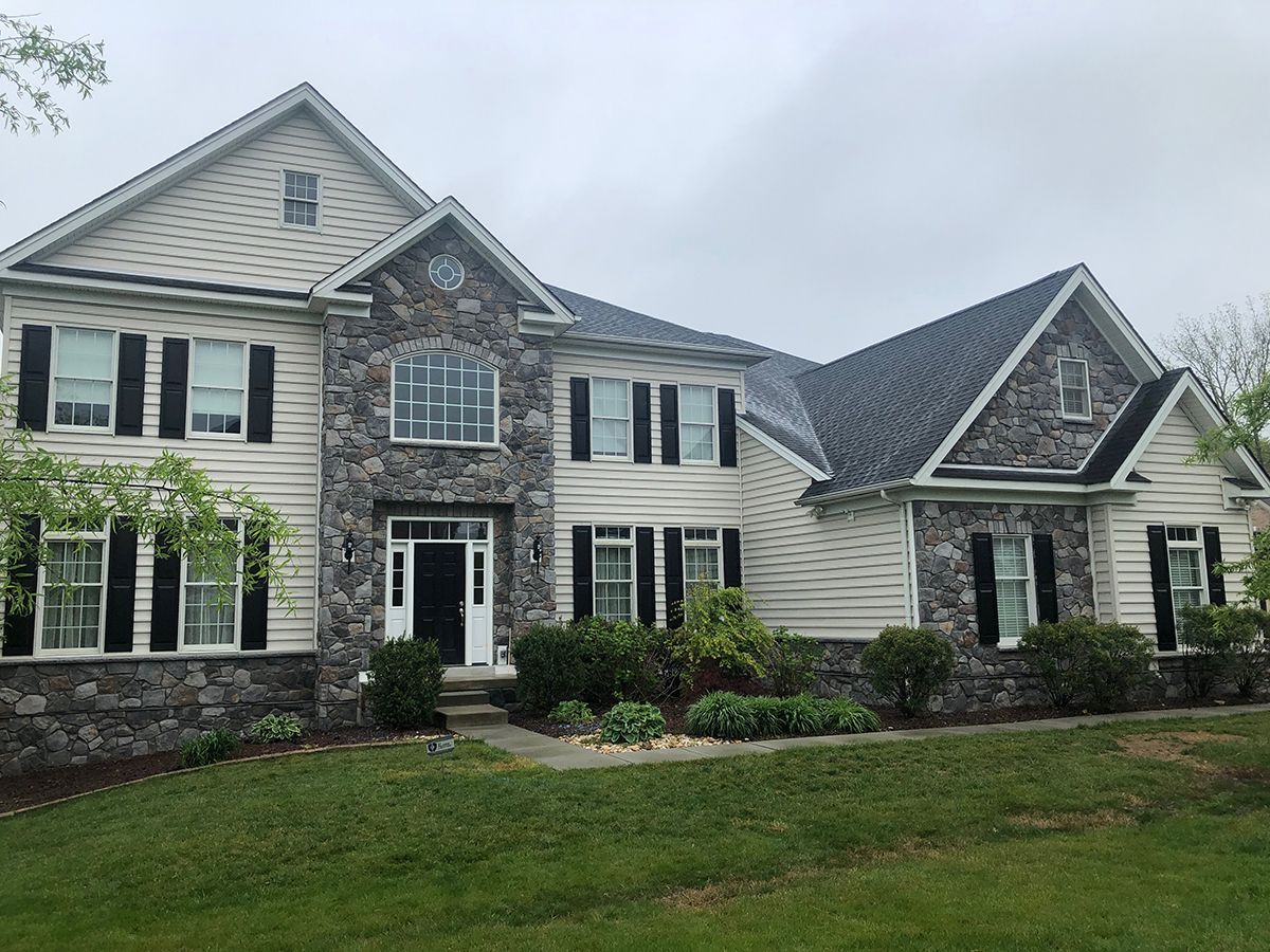 Two-story house with stone facade, beige siding, black shutters, and a dark shingled roof, set on a green lawn.