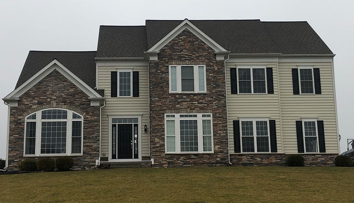 Two-story house with stone and beige siding, black shutters, and a dark roof on a cloudy day.