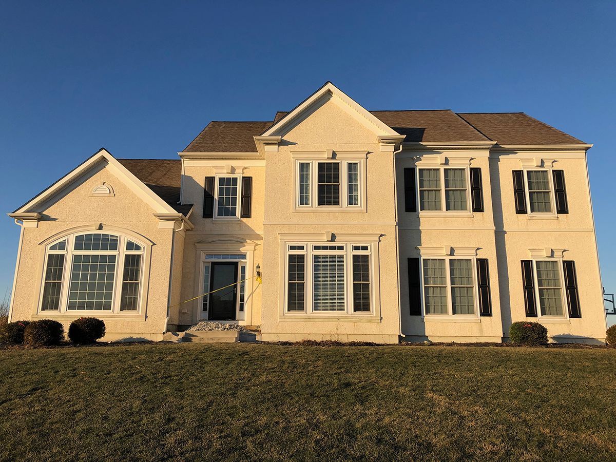 Beige two-story house with dark shutters, front door, and a brown roof against a clear blue sky.