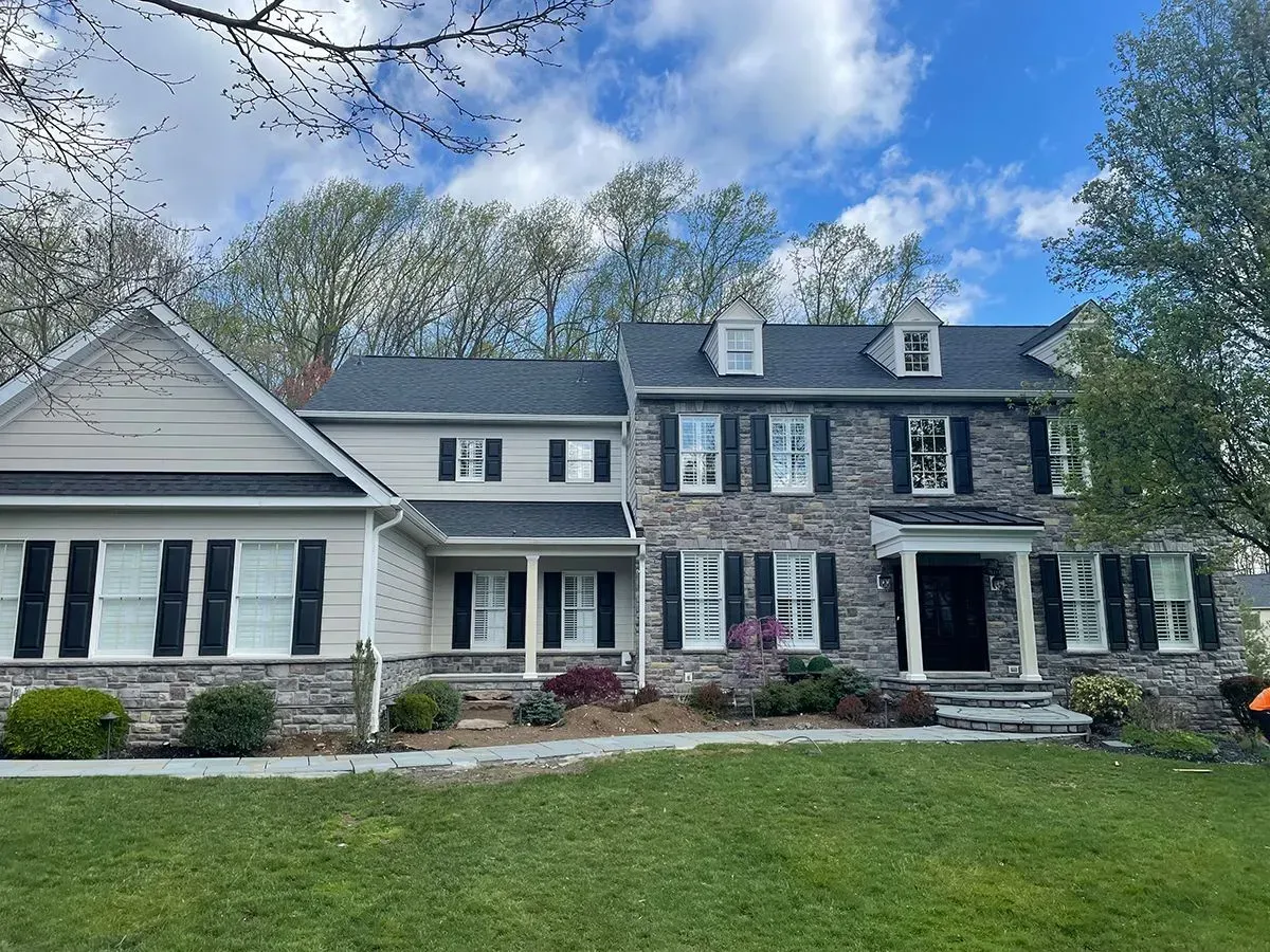 Stone-fronted house with gray roof, black shutters, and green lawn under a partly cloudy sky.