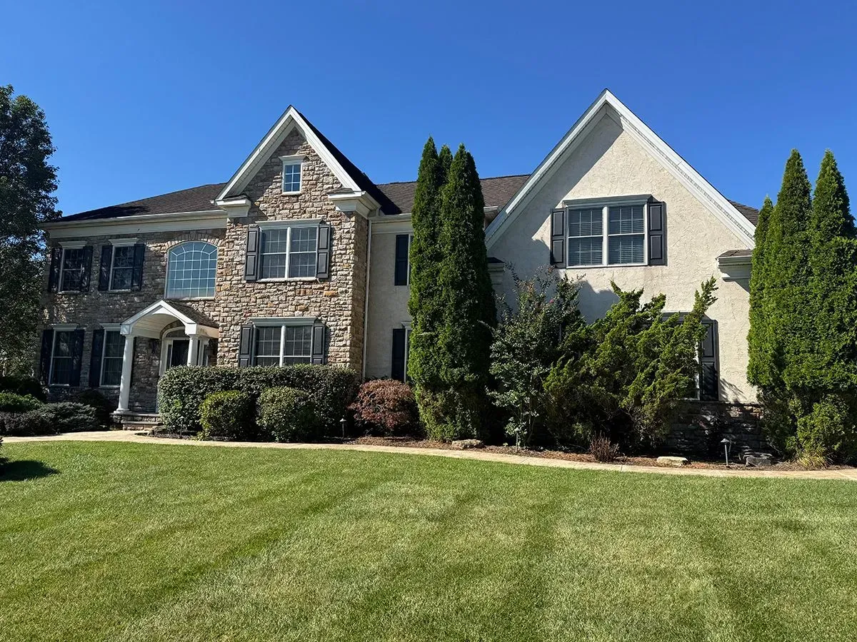 Large two-story house with stone and stucco exterior, black shutters, and well-manicured lawn under a blue sky.