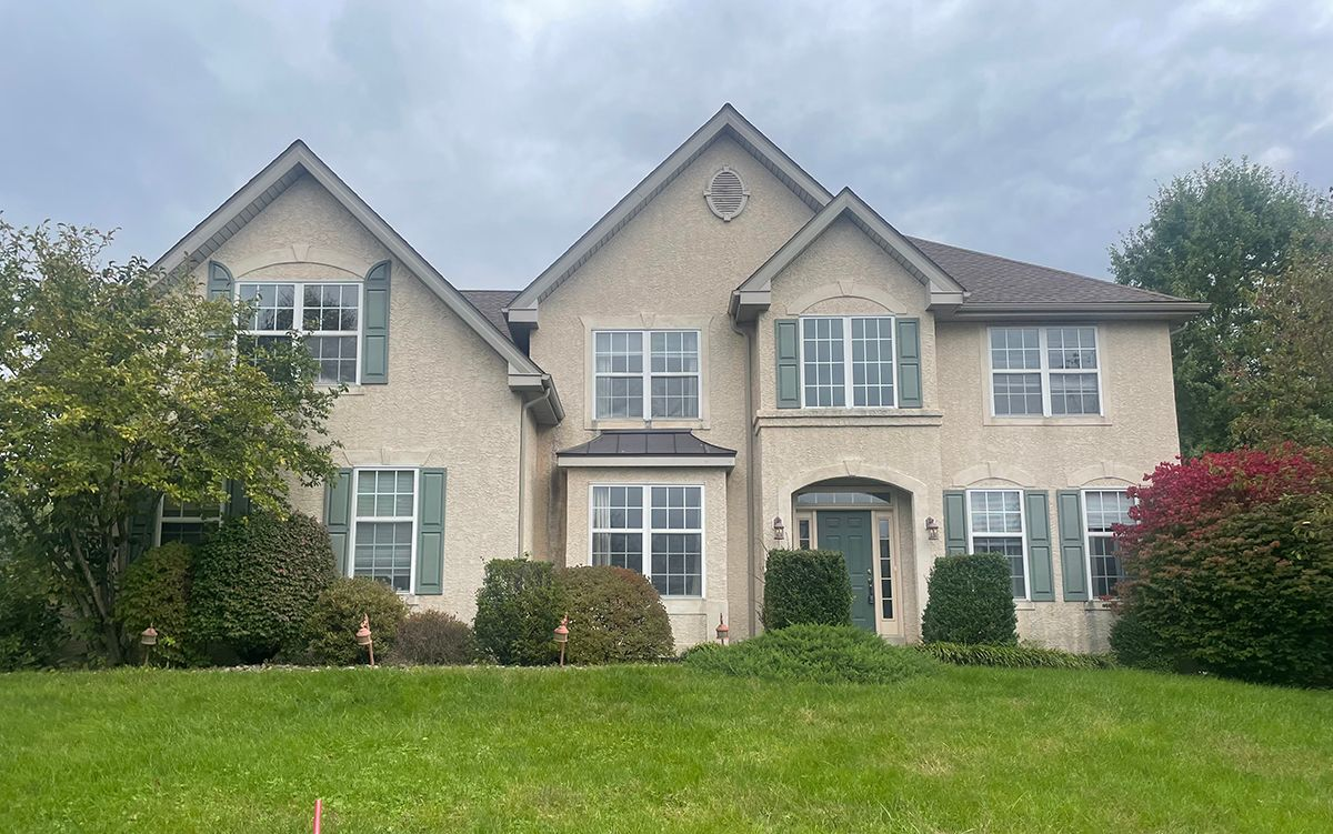 Two-story beige house with green shutters, front lawn, and cloudy sky.