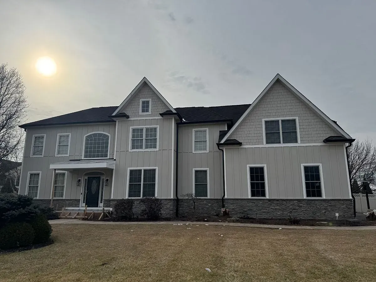 Two-story house with light siding and dark roof, gray stone base, and multiple windows on a cloudy day.