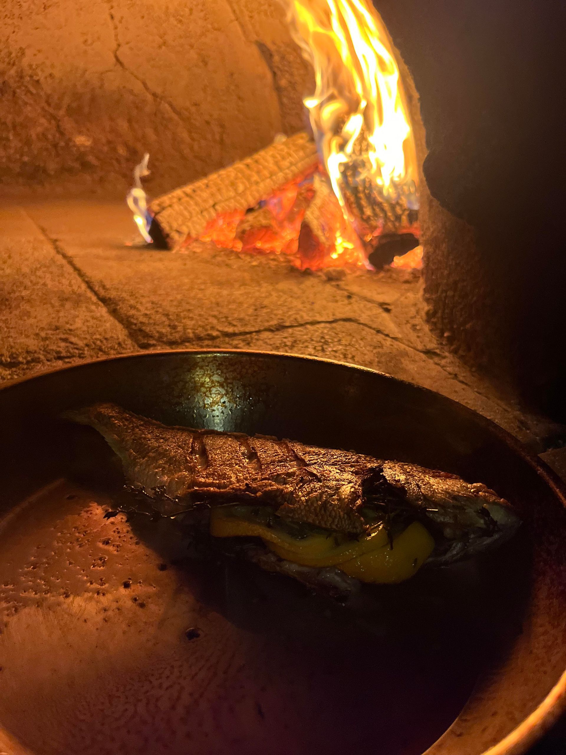 A piece of meat is cooking in a pan in front of a fire.