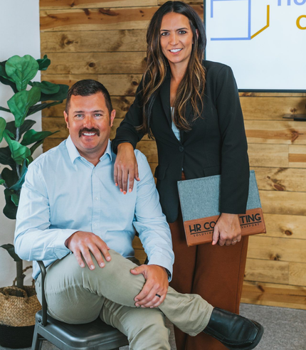 Two coworkers posing in an office lobby, one seated and one standing, both smiling.