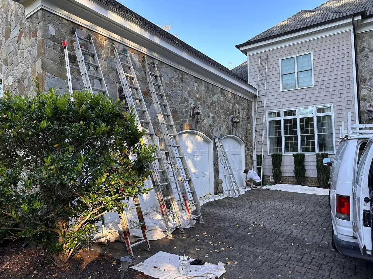 Ladders lean against a stone house, parked van nearby, likely exterior work in progress.