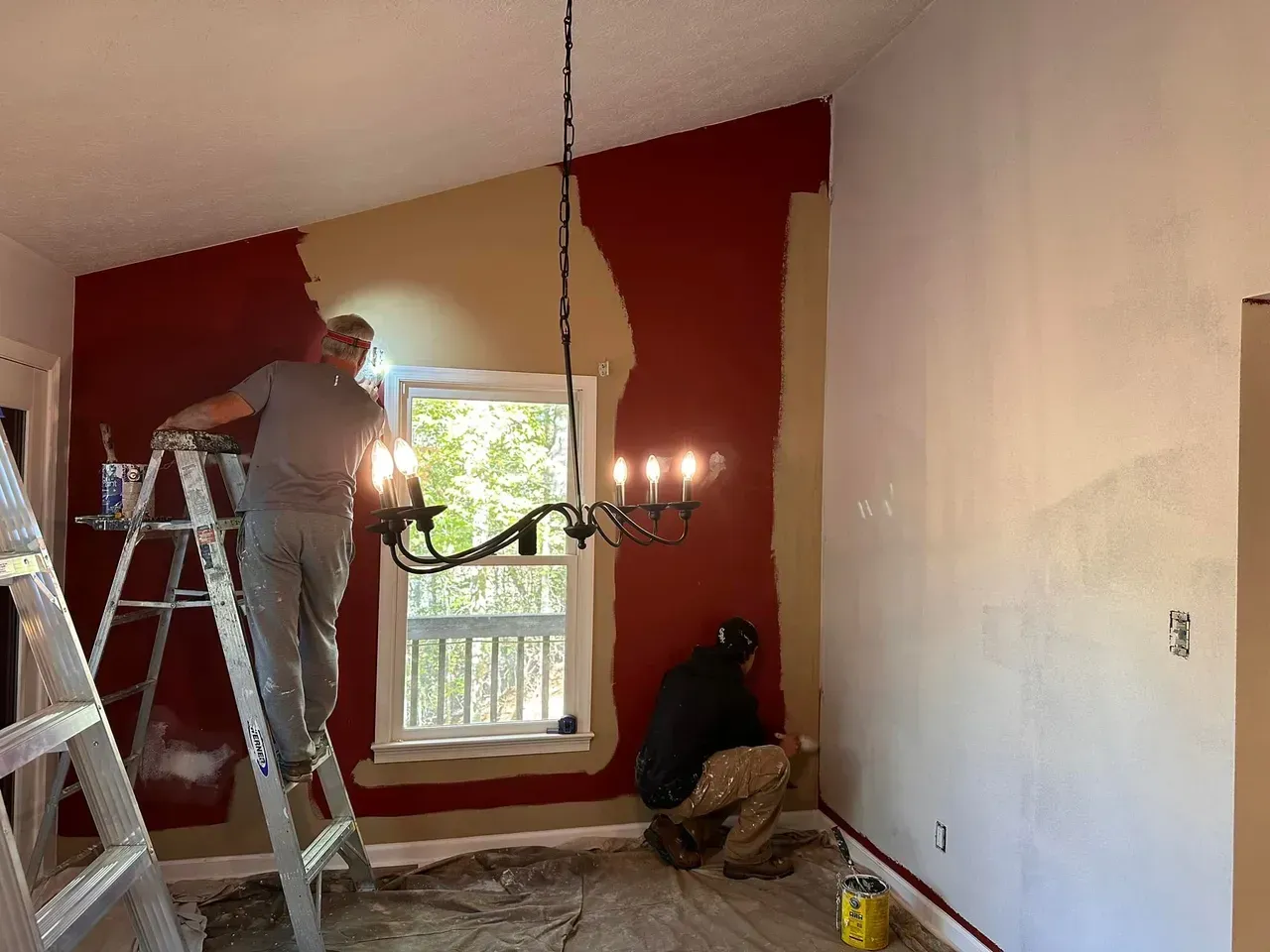 Two people paint a room: one on a ladder, the other kneeling. Red and white walls surround a window and chandelier.