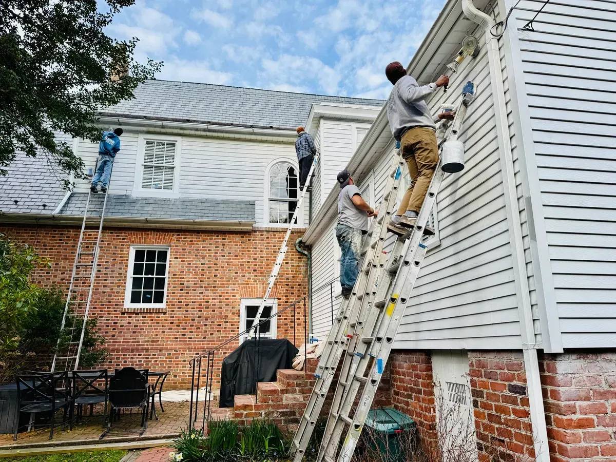 Men on ladders painting a two-story white house with a brick base.