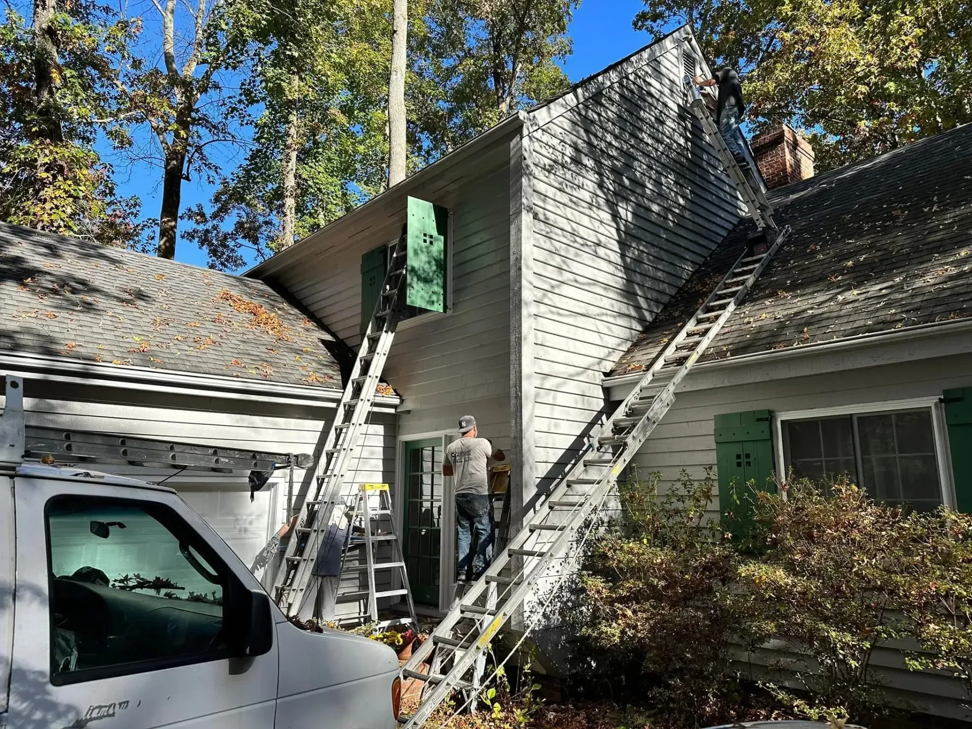 Ladder leans to a roof edge near a chimney on a house, with a truck and fall protection gear below.