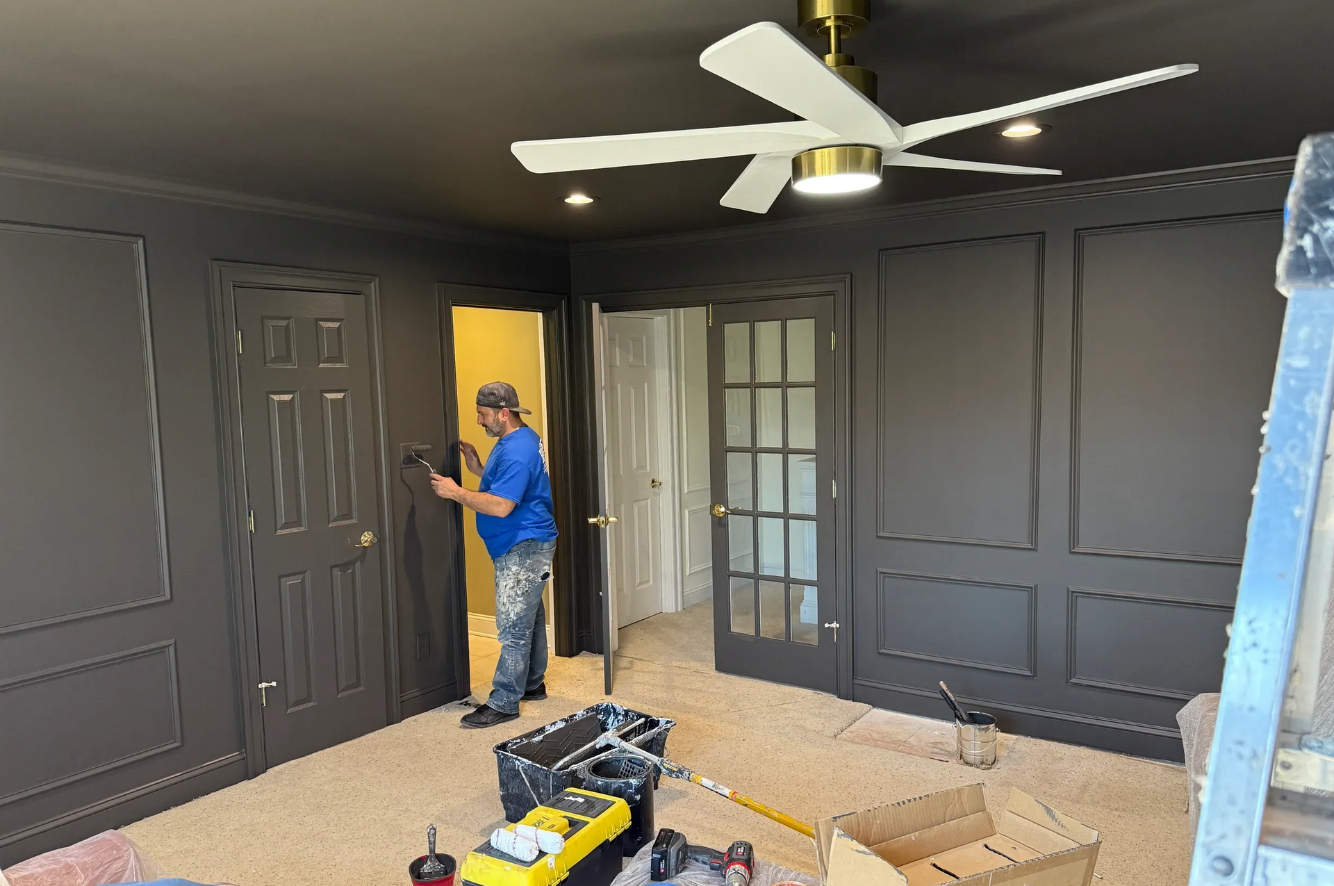 Person painting dark gray walls in a room with a white ceiling fan and open doorway.