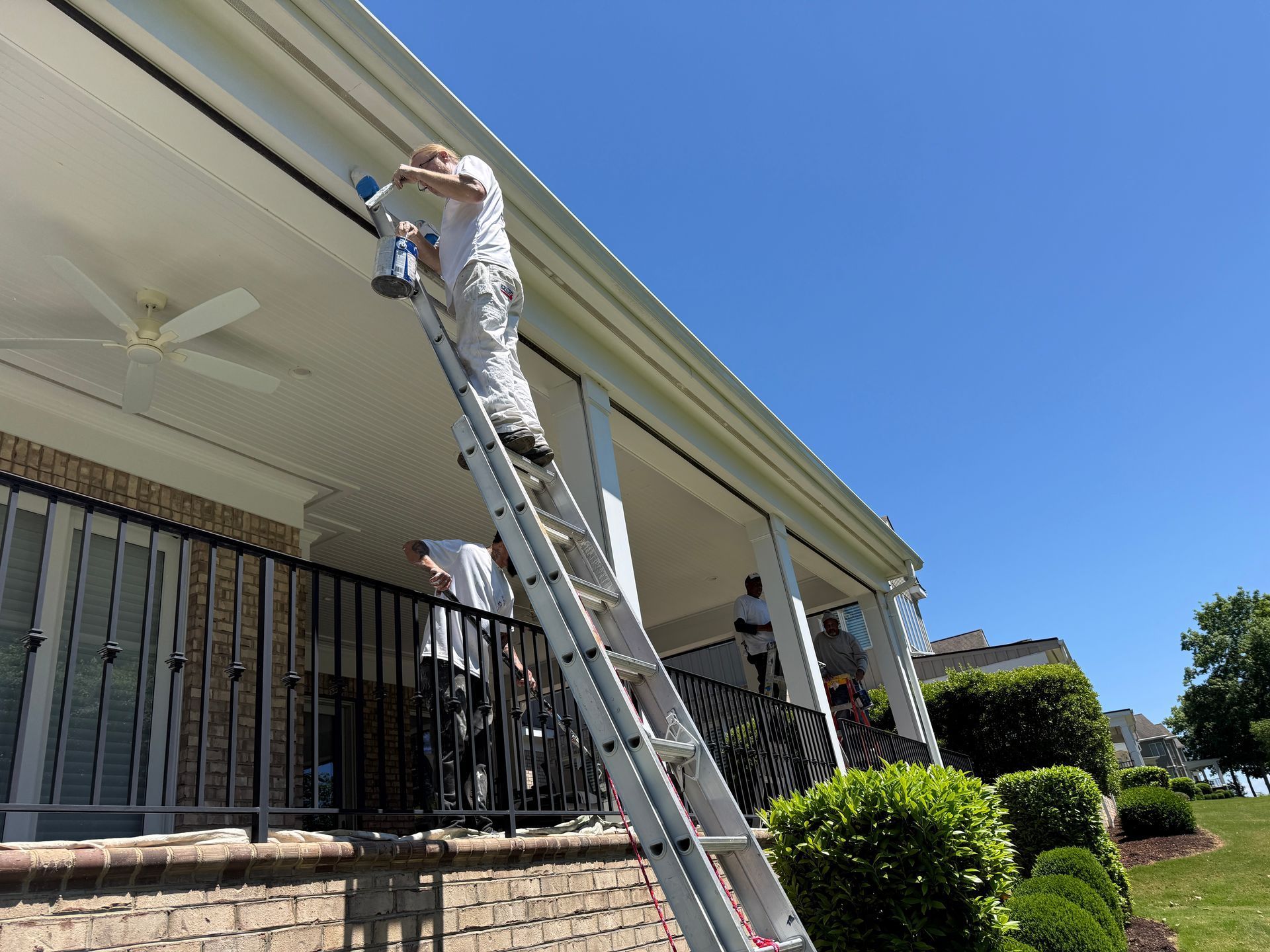 Person on a ladder painting a house’s eaves under a bright blue sky