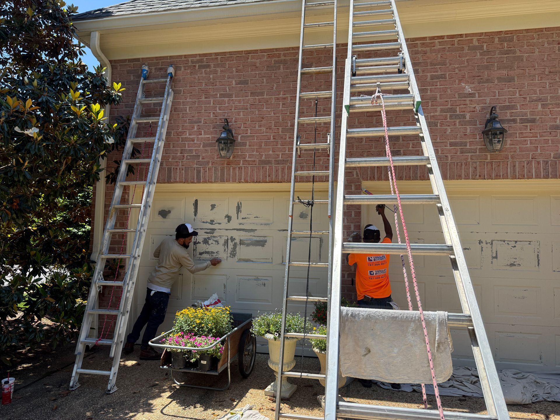 Two people on ladders painting a brick house exterior and garage doors under a porch roof
