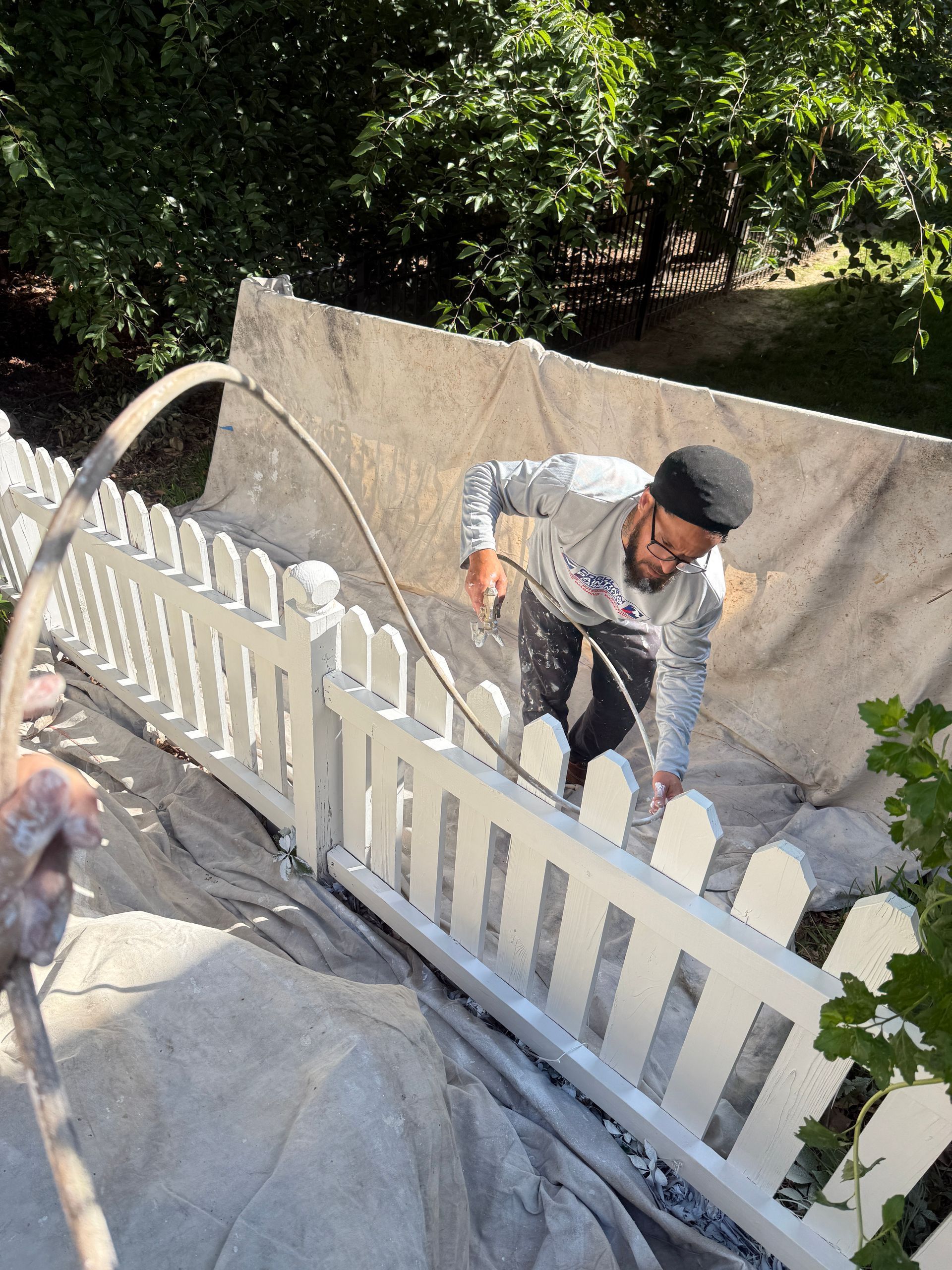 Worker painting a white fence beside a concrete wall outdoors in bright sunlight