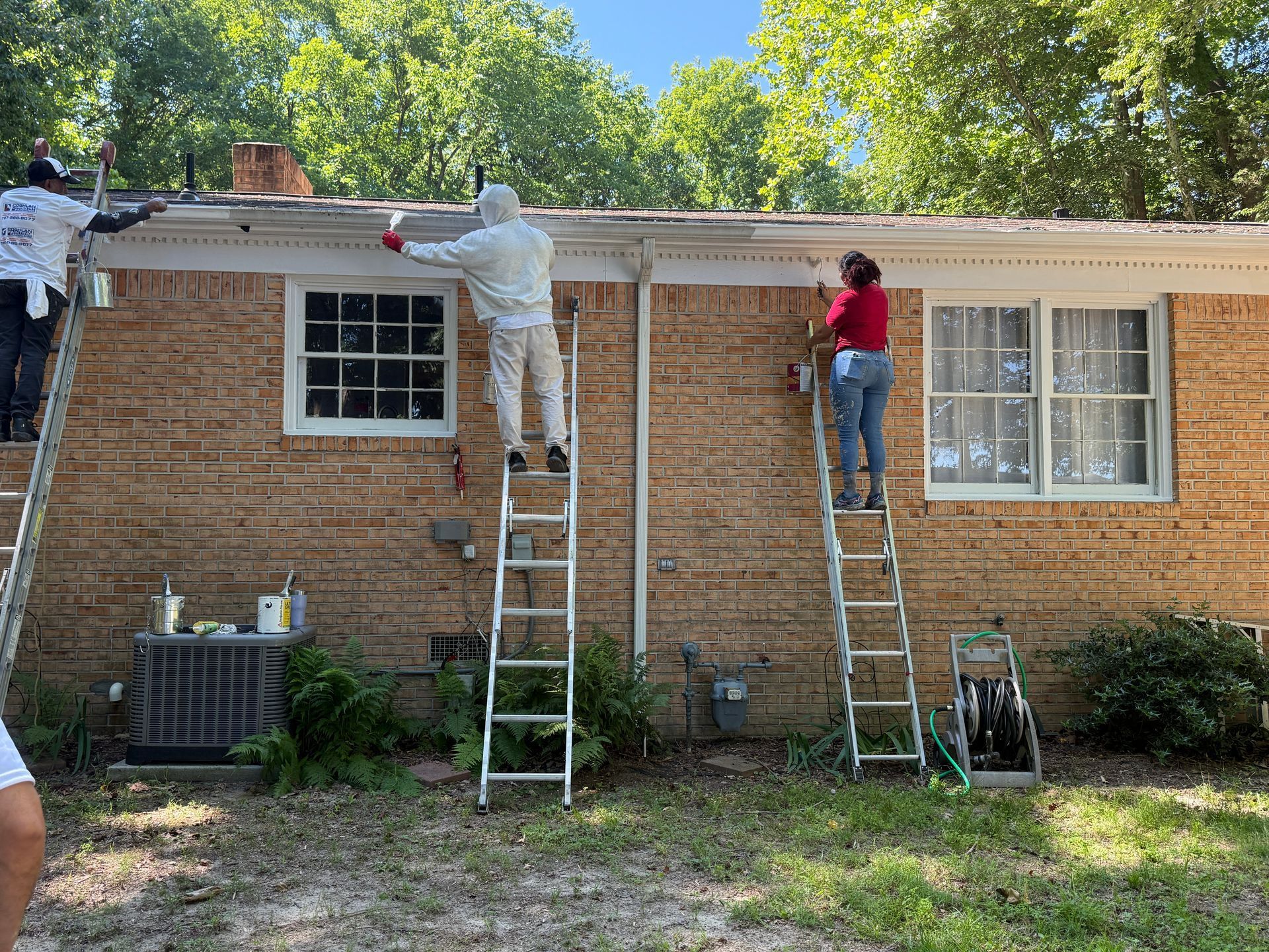 Two workers on ladders repairing a brick house roofline, with tools and buckets nearby.