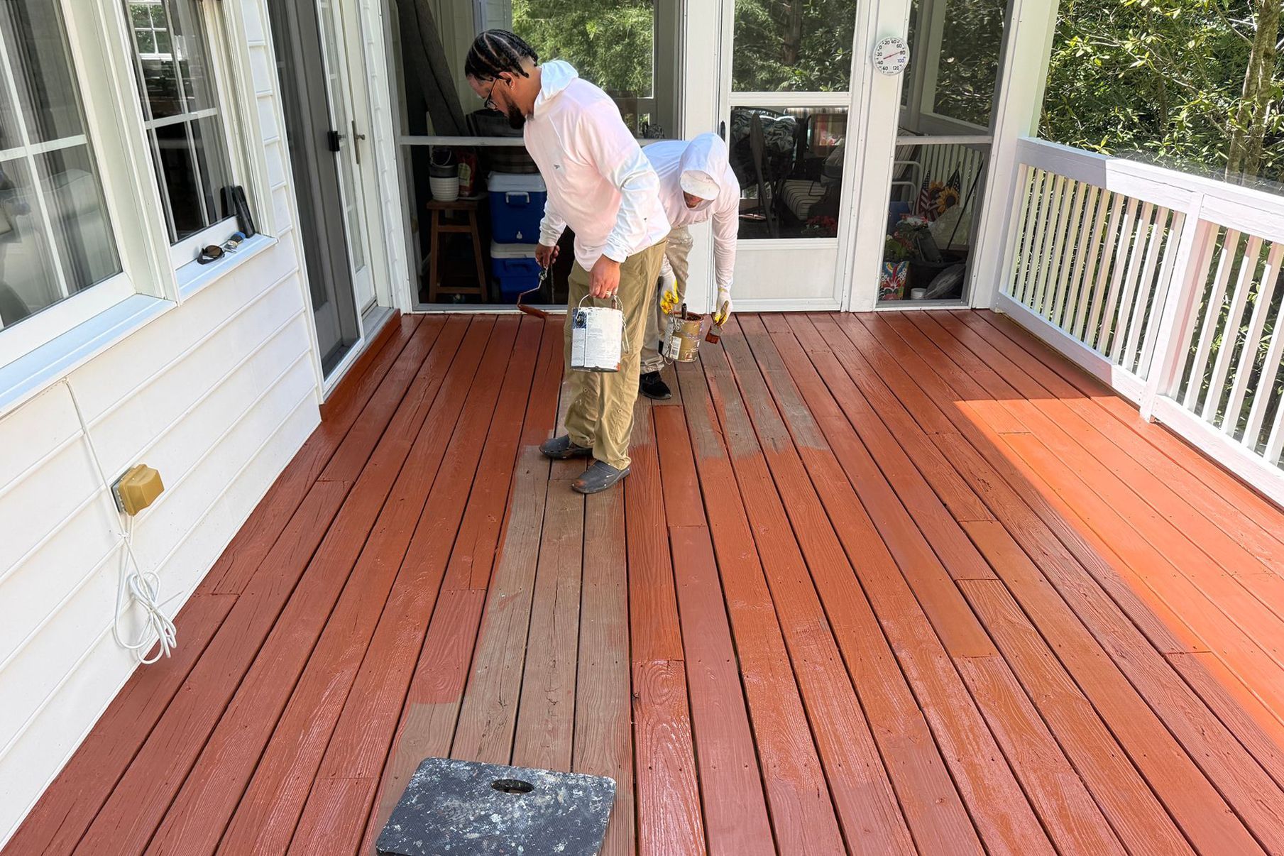 Two people staining a wooden porch deck with a paint roller and bucket