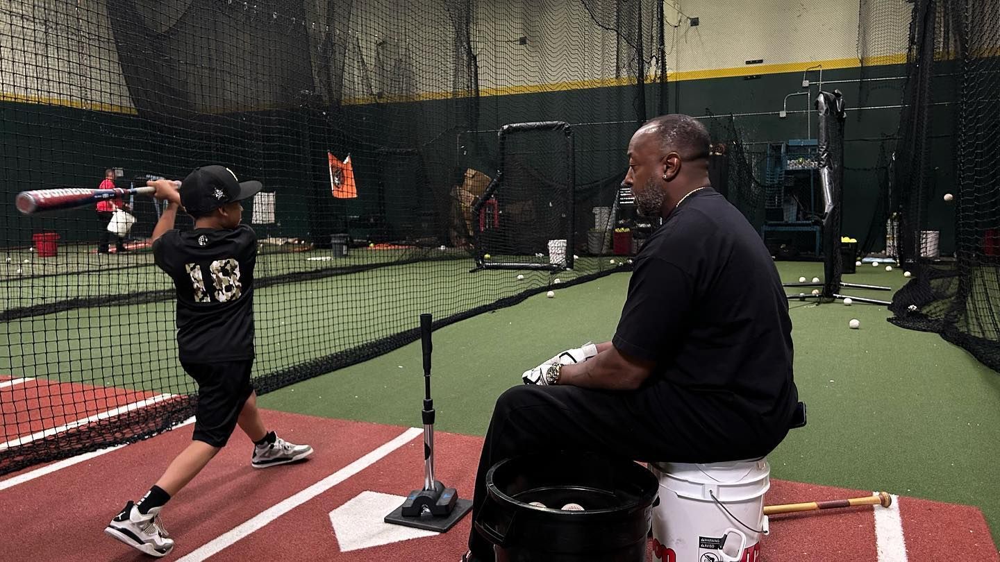 A man is sitting on a bucket watching a young boy swing a bat.