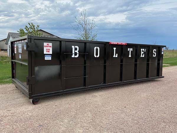 Large red dumpster filled with black trash bags in a grassy outdoor area near trees