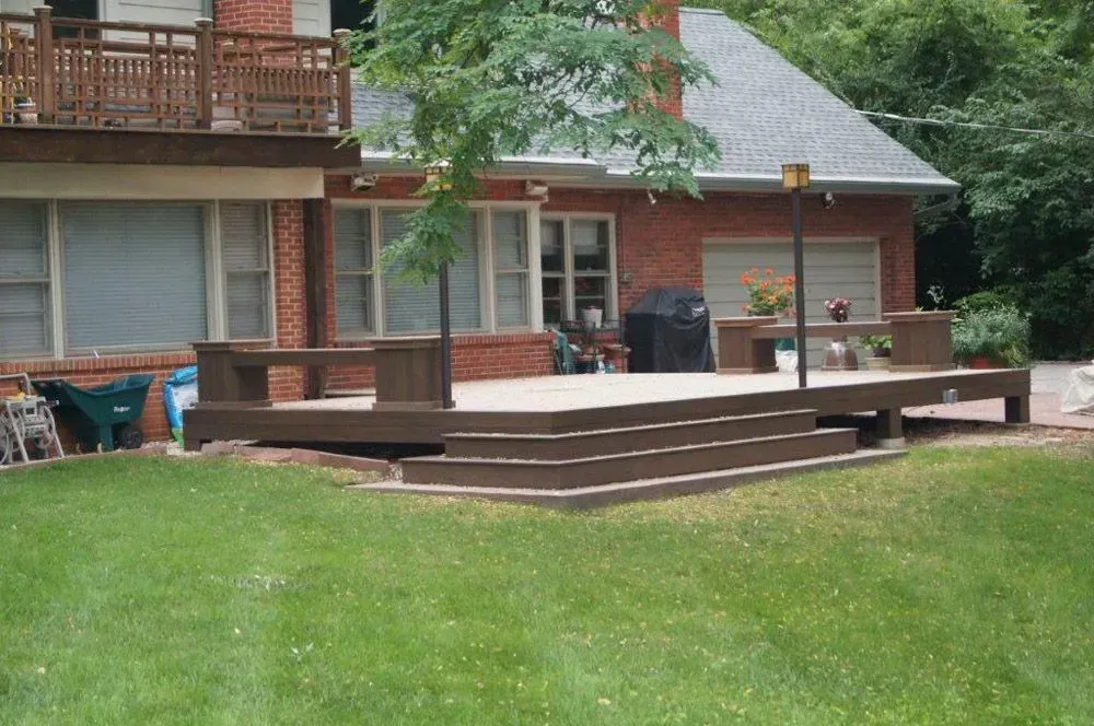 Brown wooden deck with steps, benches, and a small tree, attached to a brick house.