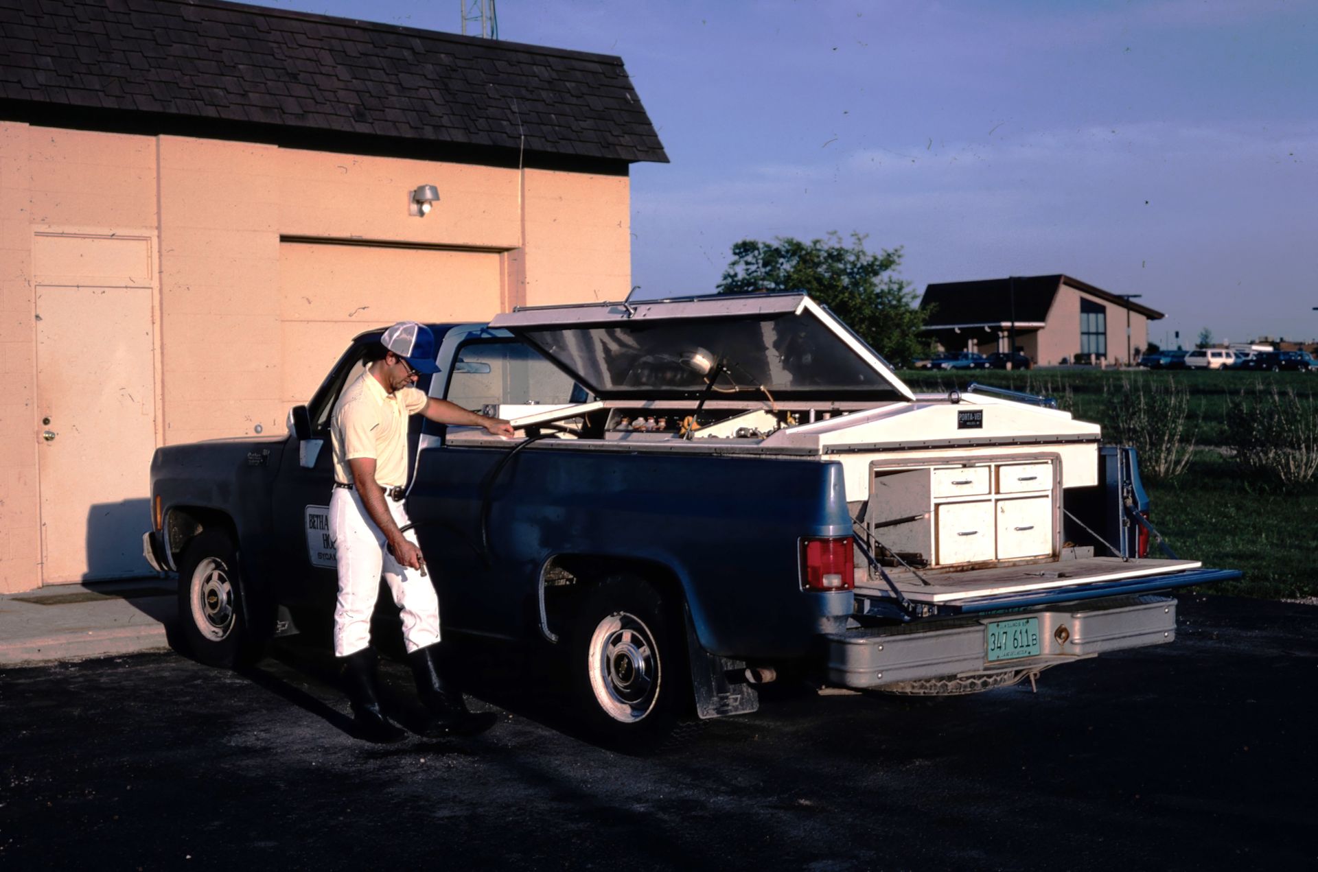 Person loading a blue pickup truck with white boxes beside a peach building at dusk