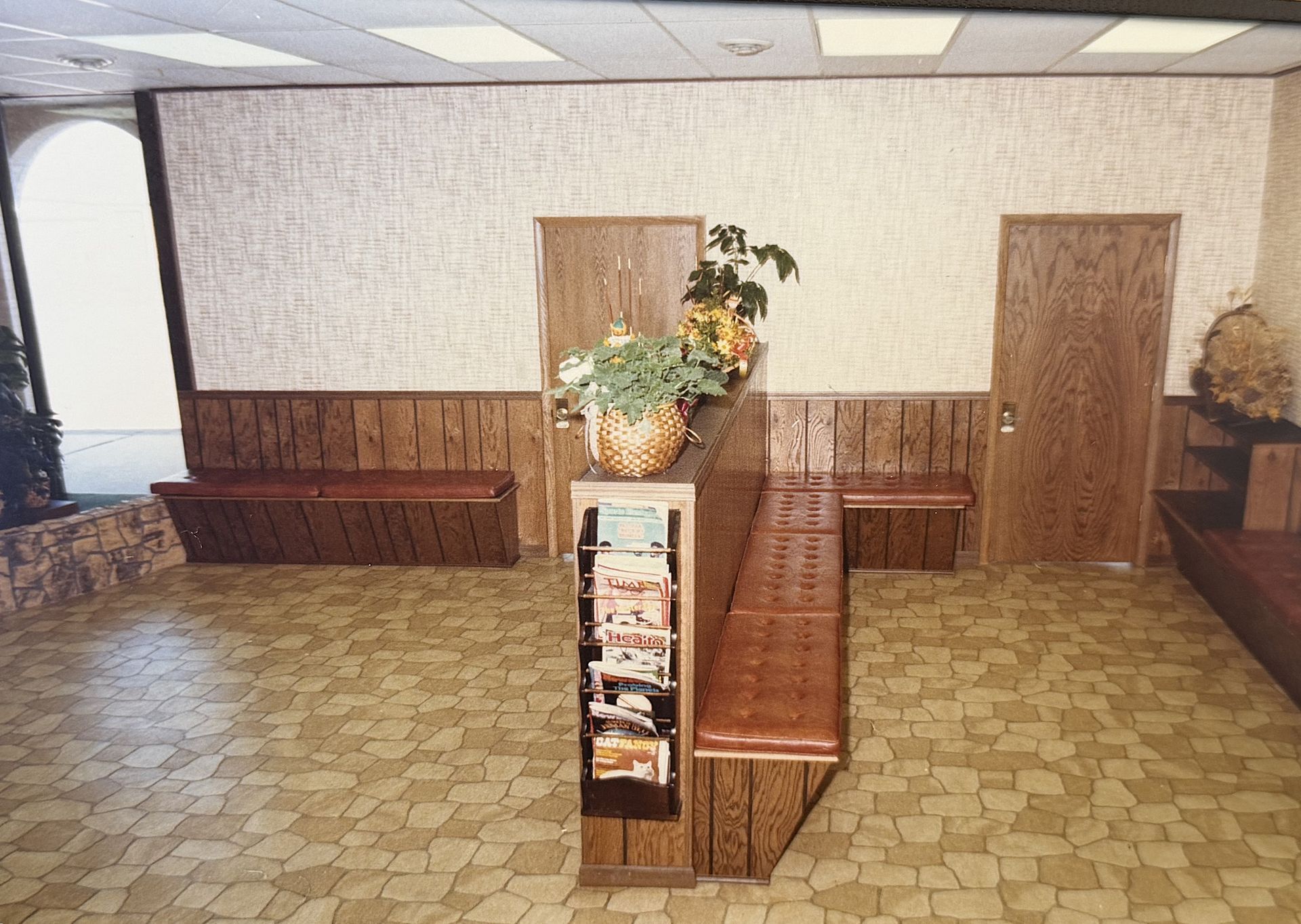 Empty waiting room with patterned carpet, wooden benches, and a central brochure stand with flowers.