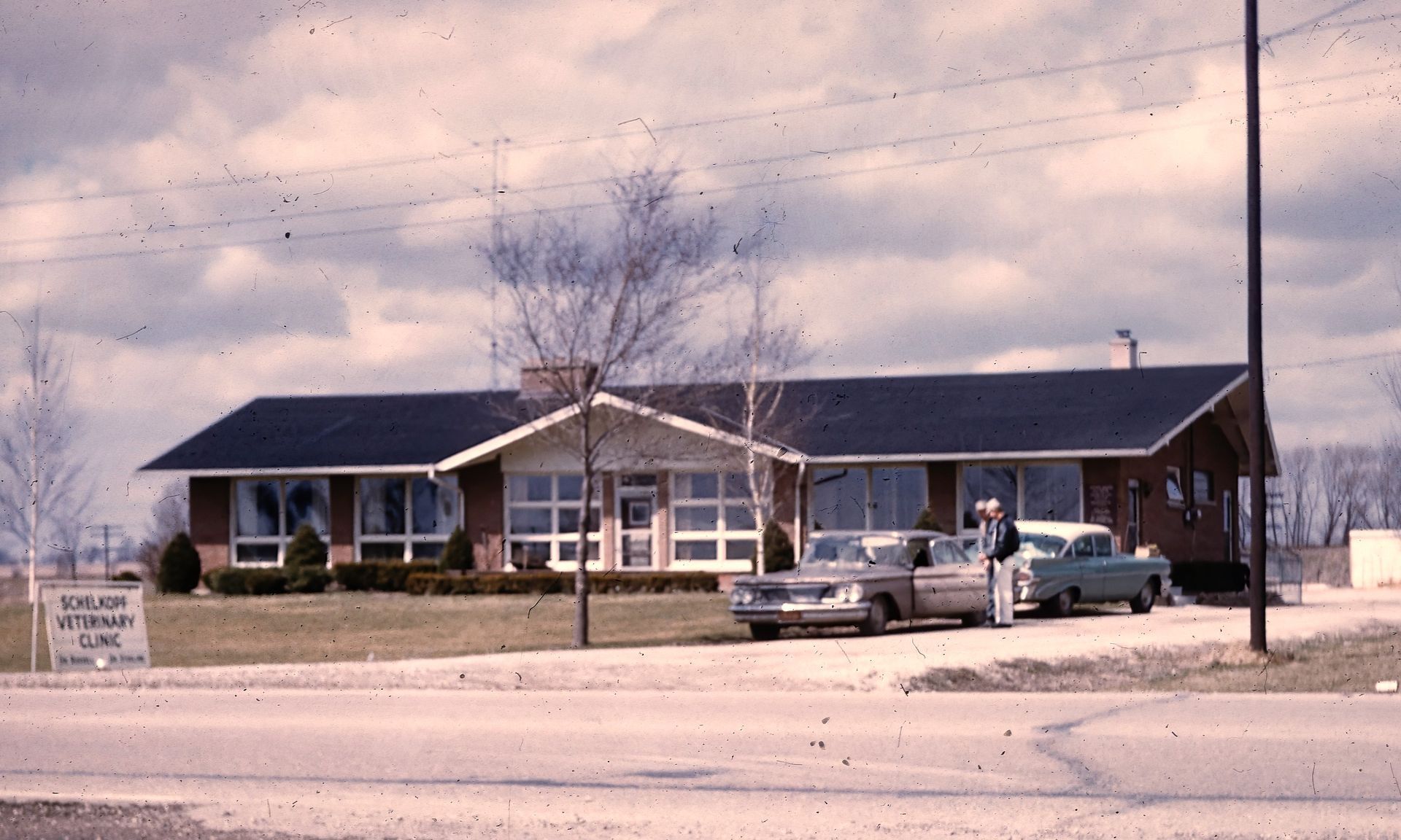 Small roadside building with a dark roof, parked cars, and a wintery sky in front.