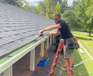 A man on a ladder repairs a gutter on a roof, surrounded by trees.