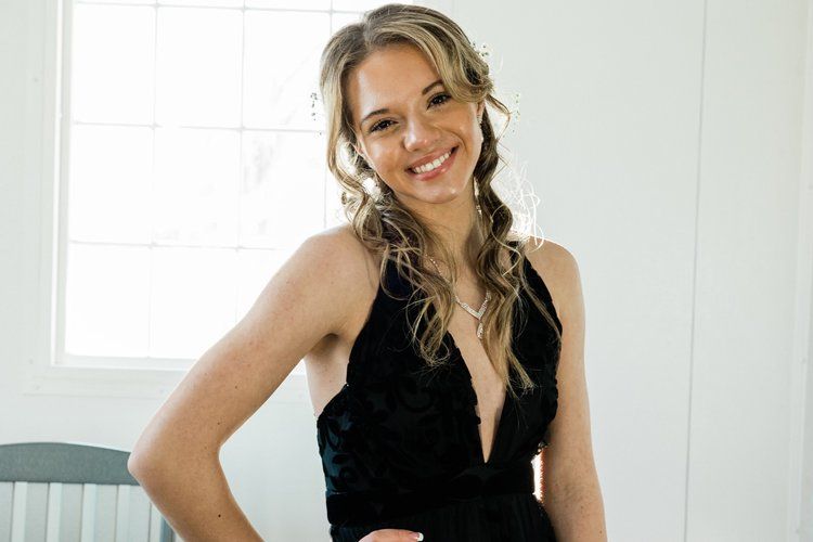 A woman in a black dress is standing in front of a window and smiling.