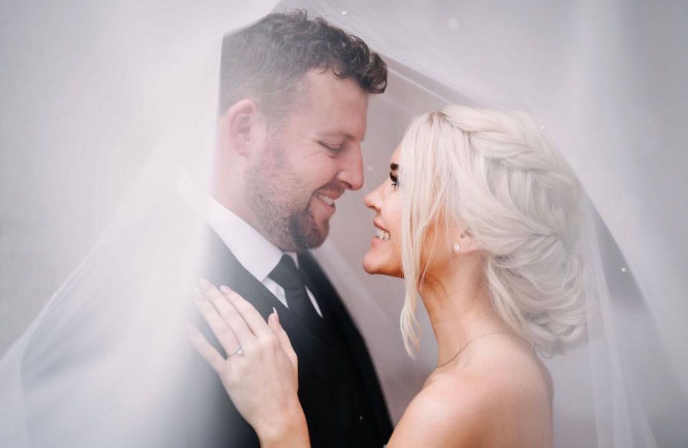 A bride and groom are standing under a veil and looking at each other.