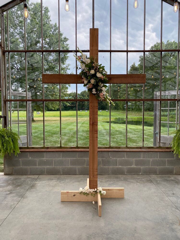 A wooden cross with flowers on it is in front of a greenhouse.