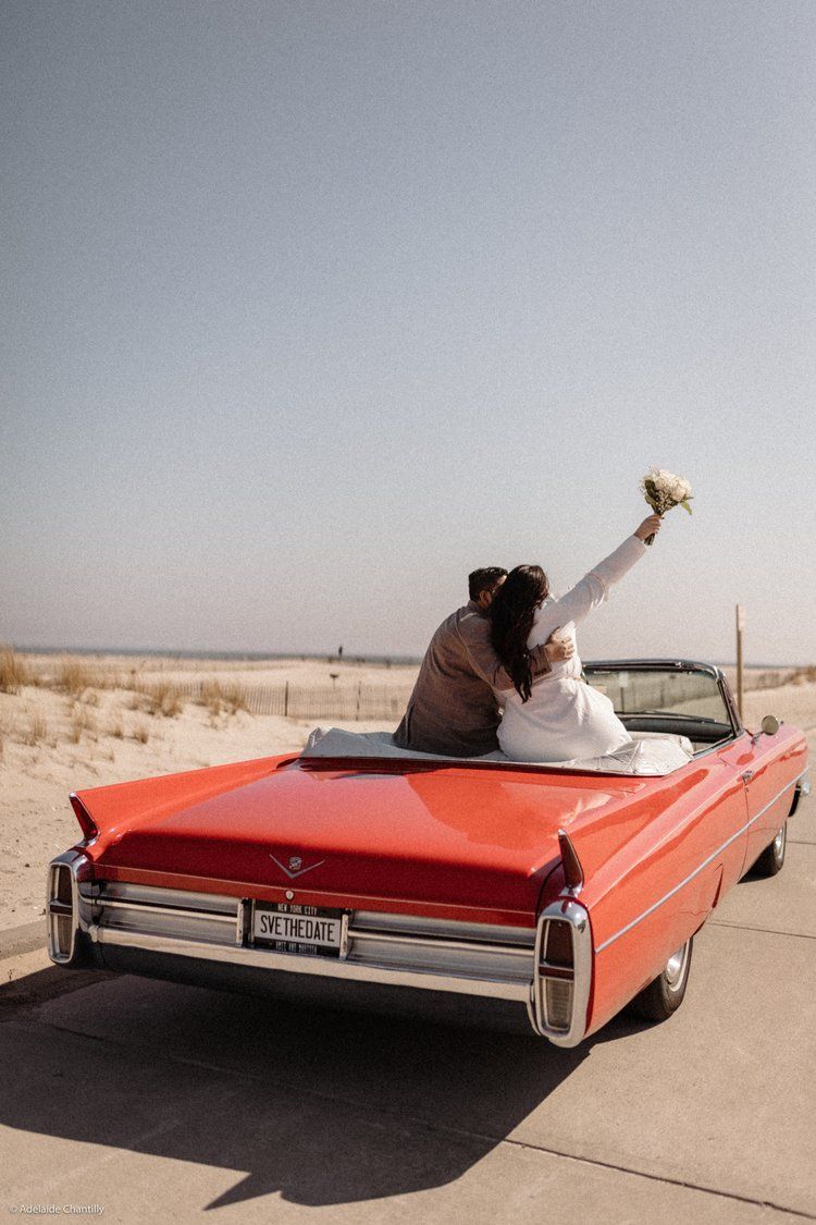 A bride and groom are sitting in the back of a red convertible car.