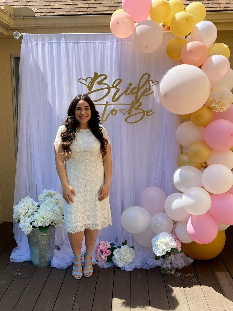 A woman in a white dress is standing in front of a bride to be backdrop.