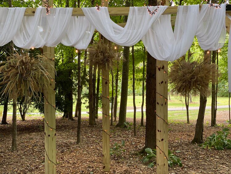 A wooden archway in the middle of a forest with white cloth hanging from it.