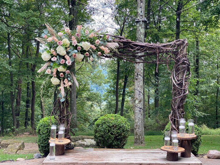 A wooden arch decorated with flowers and candles in the middle of a forest.