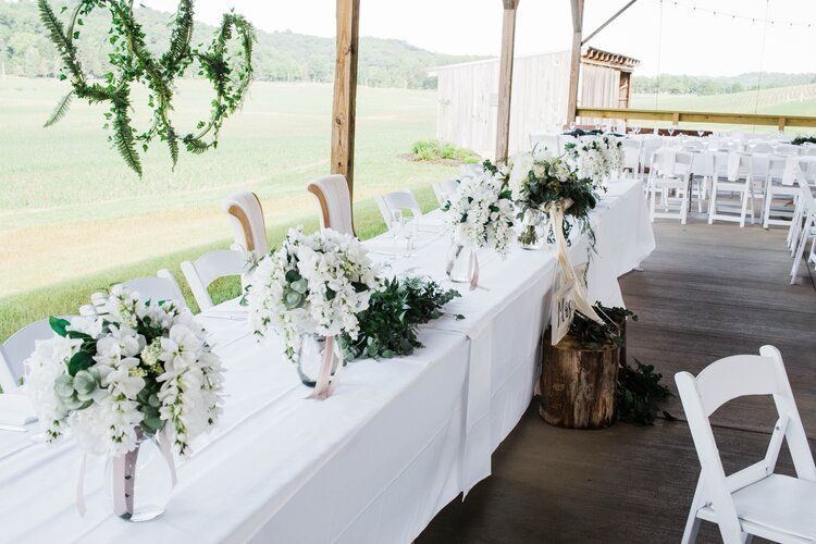 A long table with white flowers on it and white chairs.