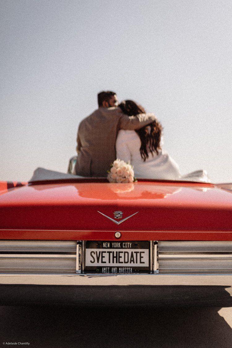 A bride and groom are sitting in the back of a red car with a license plate that says sve the date