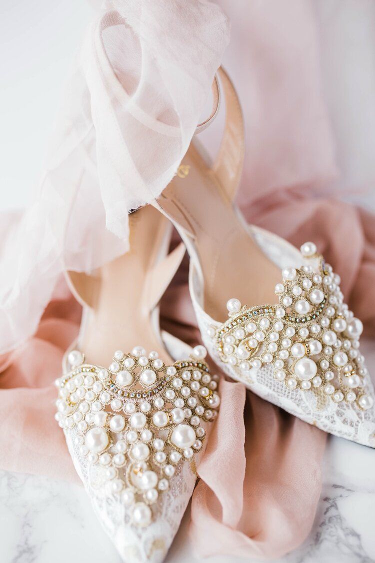 A pair of wedding shoes with pearls on them are sitting on top of a pink cloth.