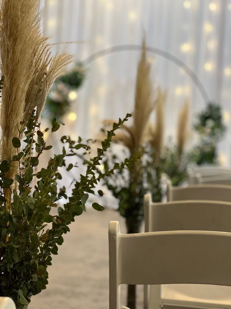A row of white chairs are lined up in a room with flowers and pampas grass.