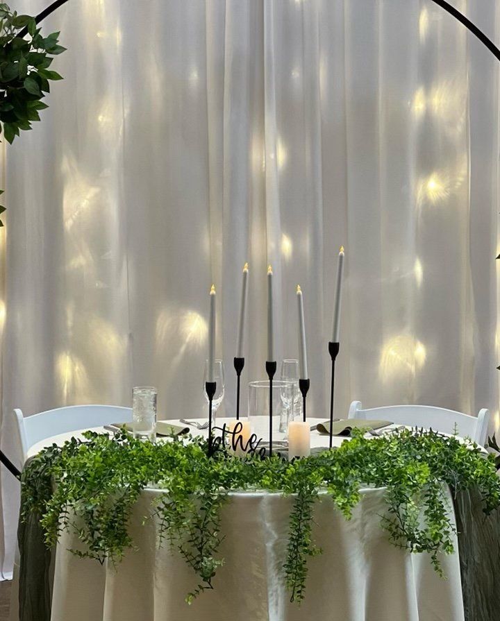 A table with candles and greenery on it in front of a white curtain.