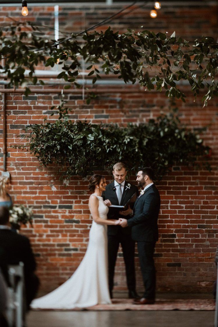 A bride and groom are holding hands during their wedding ceremony in front of a brick wall.