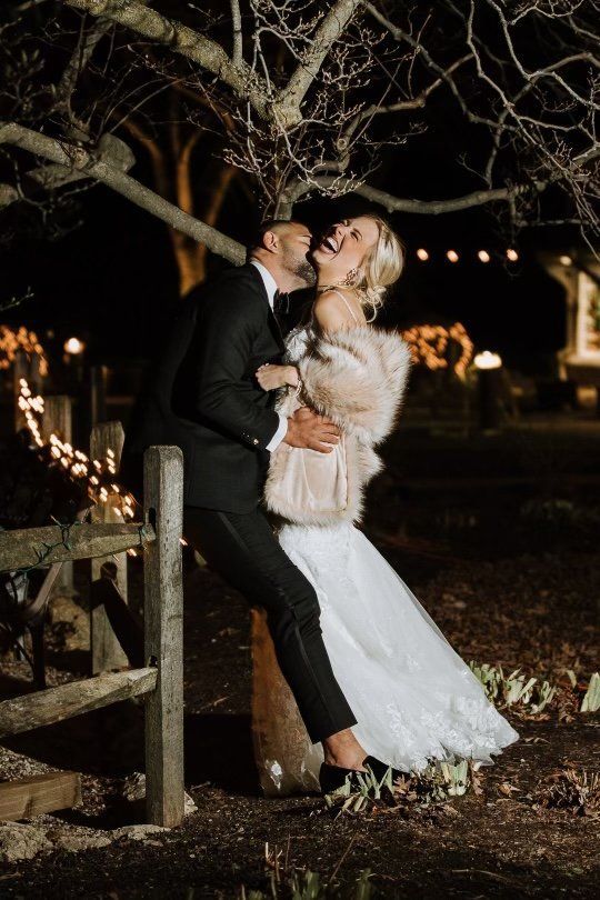 A bride and groom are leaning against a wooden fence at night.