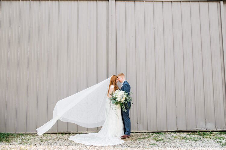 A bride and groom are posing for a picture with their veil blowing in the wind.