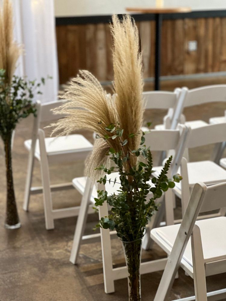 A row of white folding chairs with pampas grass in vases.