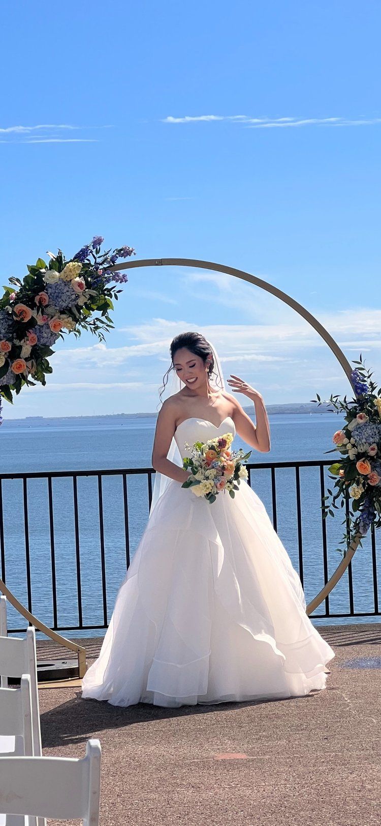 A bride in a wedding dress is standing in front of a floral arch overlooking the ocean.