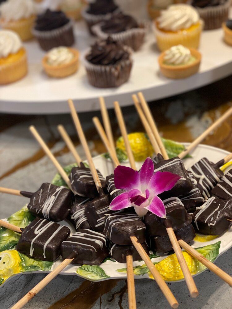 A plate of chocolate covered squares on sticks with a purple flower on top.