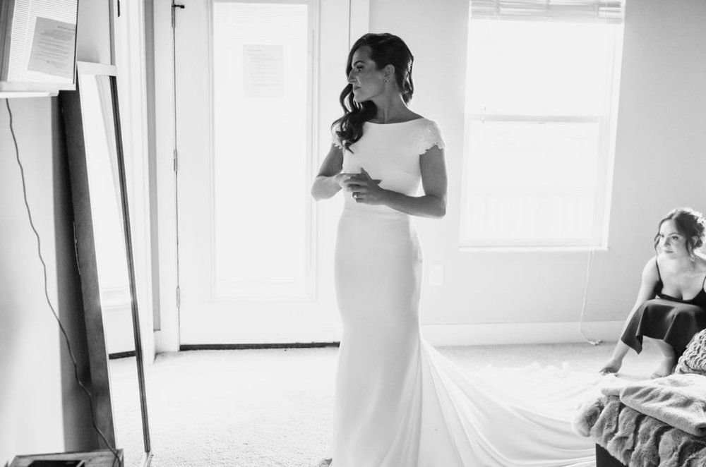 A black and white photo of a bride in a wedding dress standing in a room.