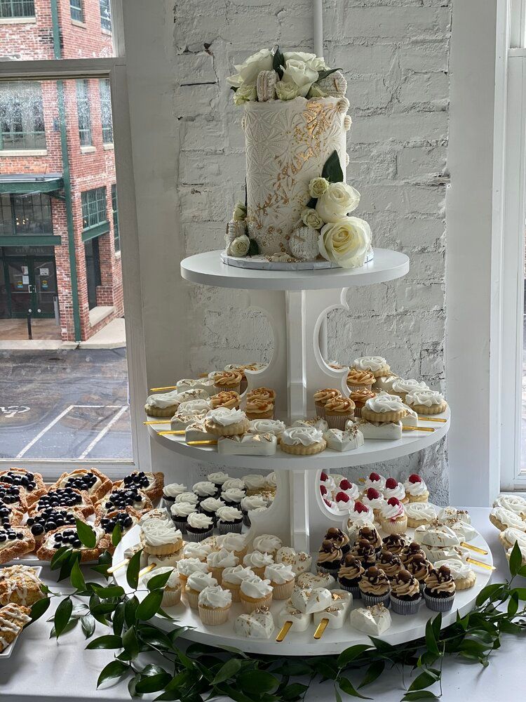 A wedding cake and cupcakes are on a table in front of a window.