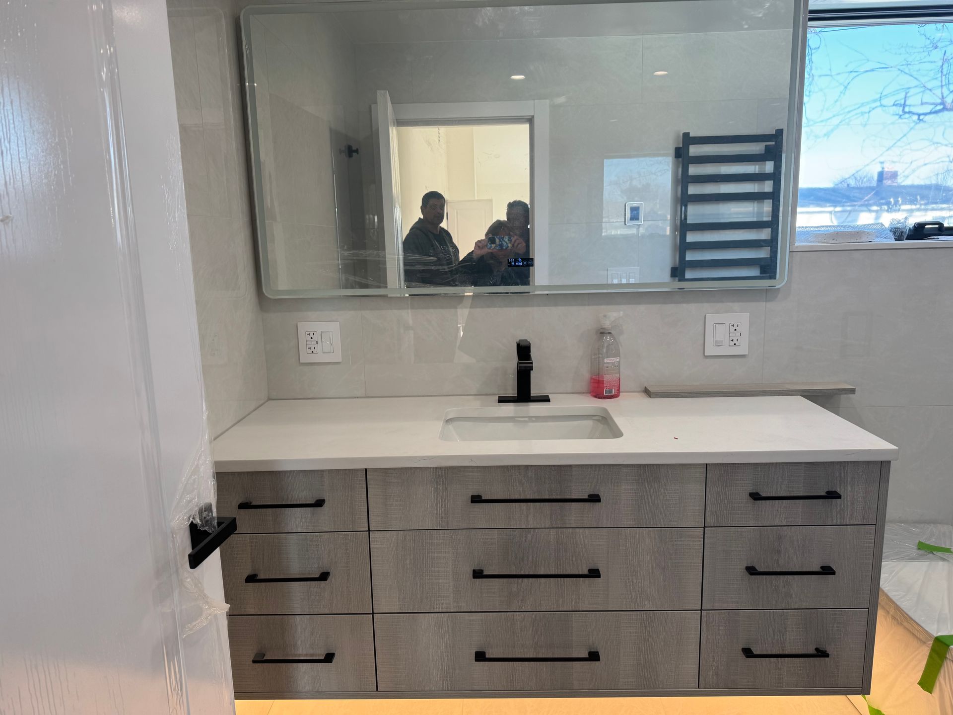 A modern bathroom vanity with gray wood-grain cabinets, black hardware, a white countertop, and a large wall mirror.